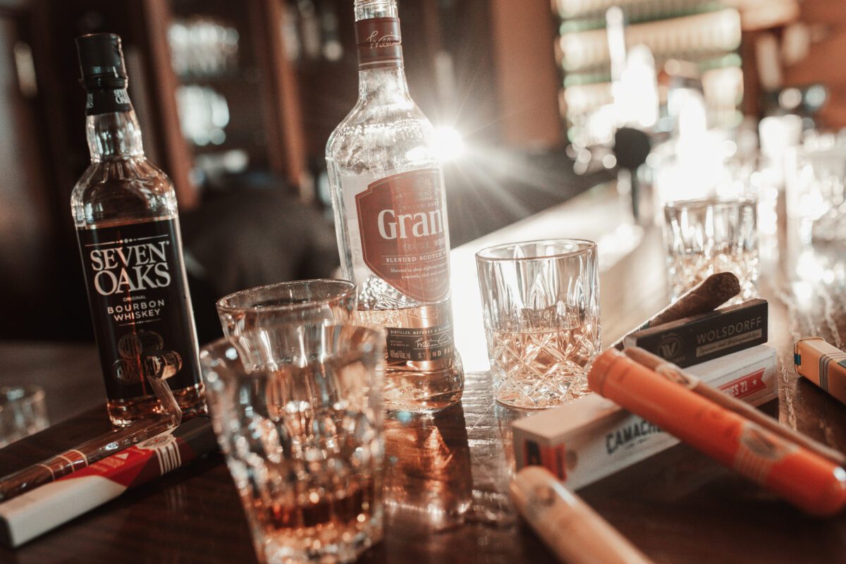 Eine Bartheke mit Flaschen Seven Oaks Bourbon Whiskey und Grand Spirits zwischen mehreren leeren Whiskeygläsern. Zahlreiche Zigarrenhülsen und ein offenes Glas mit einer Zigarre liegen verstreut herum, beleuchtet von warmem, atmosphärischem Barlicht. Die Szene ähnelt einer eleganten Kulisse, die man für einen Hochzeitsfotografen in Frankfurt finden könnte, mit Regalen voller Spirituosen im unscharfen Hintergrund. - Elis Wedding - Hochzeitsfotograf Frankfurt