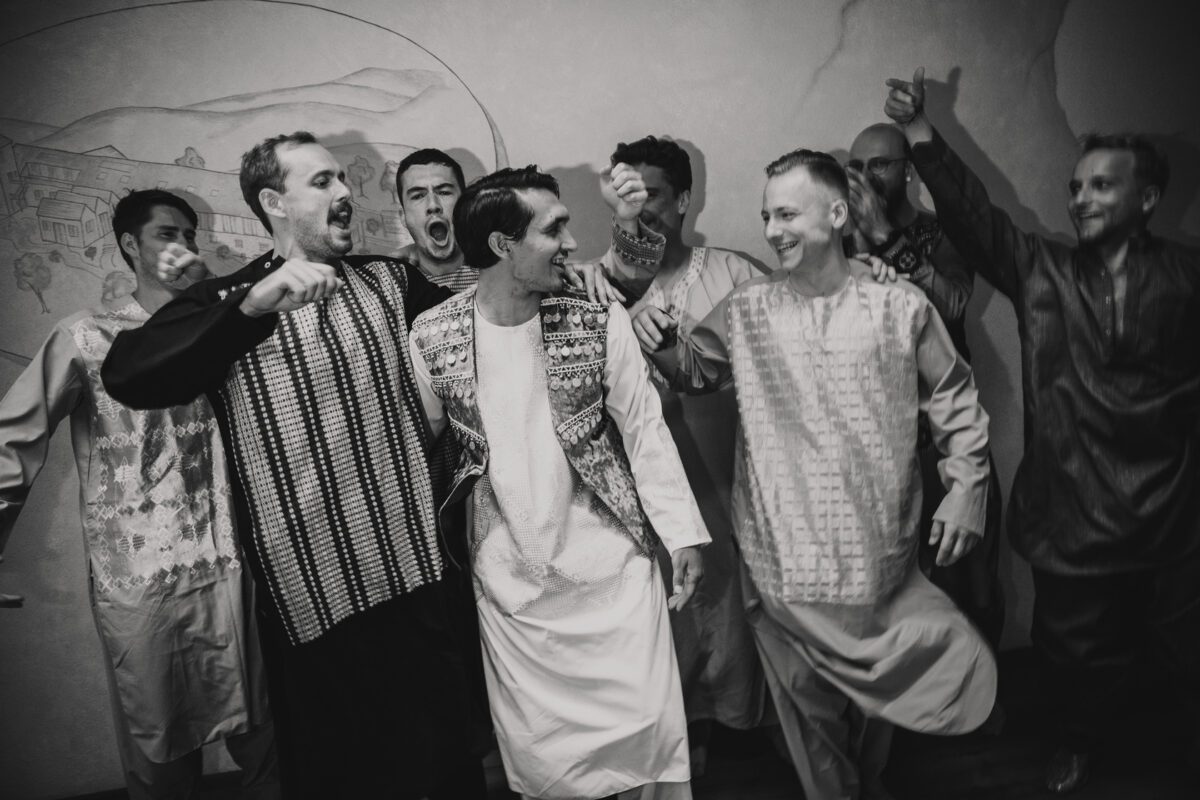 Eine Gruppe von Männern in traditioneller afghanischer Kleidung tanzt fröhlich zusammen, festgehalten in zeitlosen Schwarz-Weiß-Fotografien. Sie lächeln, haben die Arme umeinander gelegt und scheinen mitten in der Bewegung zu sein. Im Hintergrund ist ein Wandgemälde mit Bergen und Gebäuden zu sehen, das die Energie und Kameradschaft des Augenblicks einfängt. - Elis Wedding - Hochzeitsfotograf Frankfurt