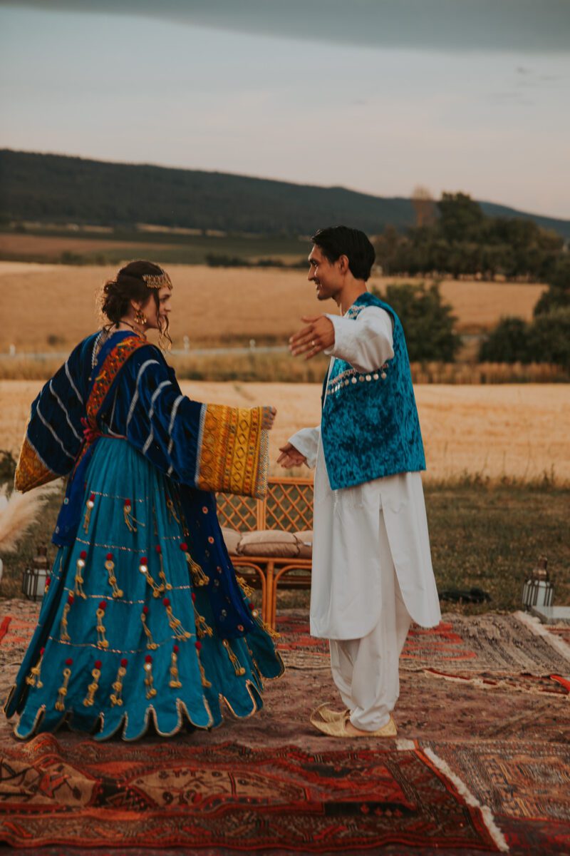 Ein Paar in traditioneller Kleidung tanzt im Freien, möglicherweise zur Feier seiner standesamtlichen Trauung. Die Frau trägt ein blaues, gemustertes Kleid mit gelben Akzenten, während der Mann ein weißes Outfit mit einer blauen Weste trägt. Sie stehen auf bunten Teppichen vor Feldern und fernen Bergen unter einem klaren Himmel. - Elis Wedding - Hochzeitsfotograf Frankfurt