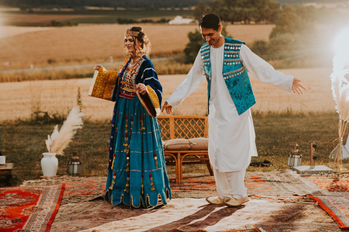 Ein Mann und eine Frau in traditioneller Kleidung tanzen im Freien auf einem farbenfrohen Teppich bei Sonnenuntergang. Die Frau trägt ein blaues, verziertes Kleid und hält Fächer, während der Mann ein weißes Outfit mit einer blauen Weste trägt. Sie wirken fröhlich, umgeben von Feldern und rustikalem Dekor. Dieser Moment wurde in wunderschönen Fotos festgehalten und strahlt Festlichkeit aus. - Elis Wedding - Hochzeitsfotograf Frankfurt