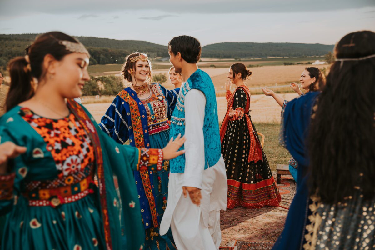 Eine Gruppe von Menschen in farbenfroher traditioneller Kleidung tanzt in einer malerischen Umgebung im Freien, wunderschön eingefangen von einem Hochzeitsfotografen. Die Frauen tragen farbenfrohe Kleider mit aufwendigen Stickereien und Stirnbändern, während die Männer weiße Outfits mit blauen Westen tragen. Offene Felder und Hügel erstrecken sich unter einem teilweise bewölkten Himmel. - Elis Wedding - Hochzeitsfotograf Frankfurt