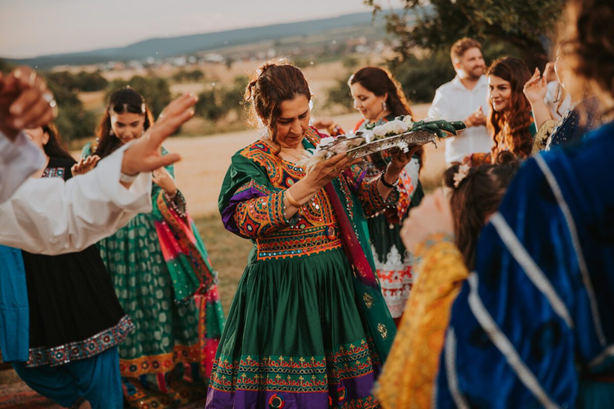 Eine Gruppe von Menschen in farbenfroher traditioneller Kleidung versammelt sich draußen auf einer Wiese unter einem teilweise bewölkten Himmel. Eine Frau in der Mitte, die ein grünes Kleid mit komplizierten Mustern trägt, hält ein großes dekoriertes Tablett. Das Fotoshooting fängt den freudigen Moment ein, in dem alle das festliche Treiben genießen. - Elis Wedding - Hochzeitsfotograf Frankfurt