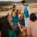 Ein Mann und eine Frau in traditioneller, farbenfroher Kleidung tanzen fröhlich bei einem Fest im Freien in der Nähe von Frankfurt. Sie sind von sitzenden Menschen umgeben, die klatschen und lächeln. Im Hintergrund ist eine malerische Landschaft mit Feldern und sanften Hügeln unter einem leicht bewölkten Himmel in der Abenddämmerung zu sehen. - Elis Wedding - Hochzeitsfotograf Frankfurt