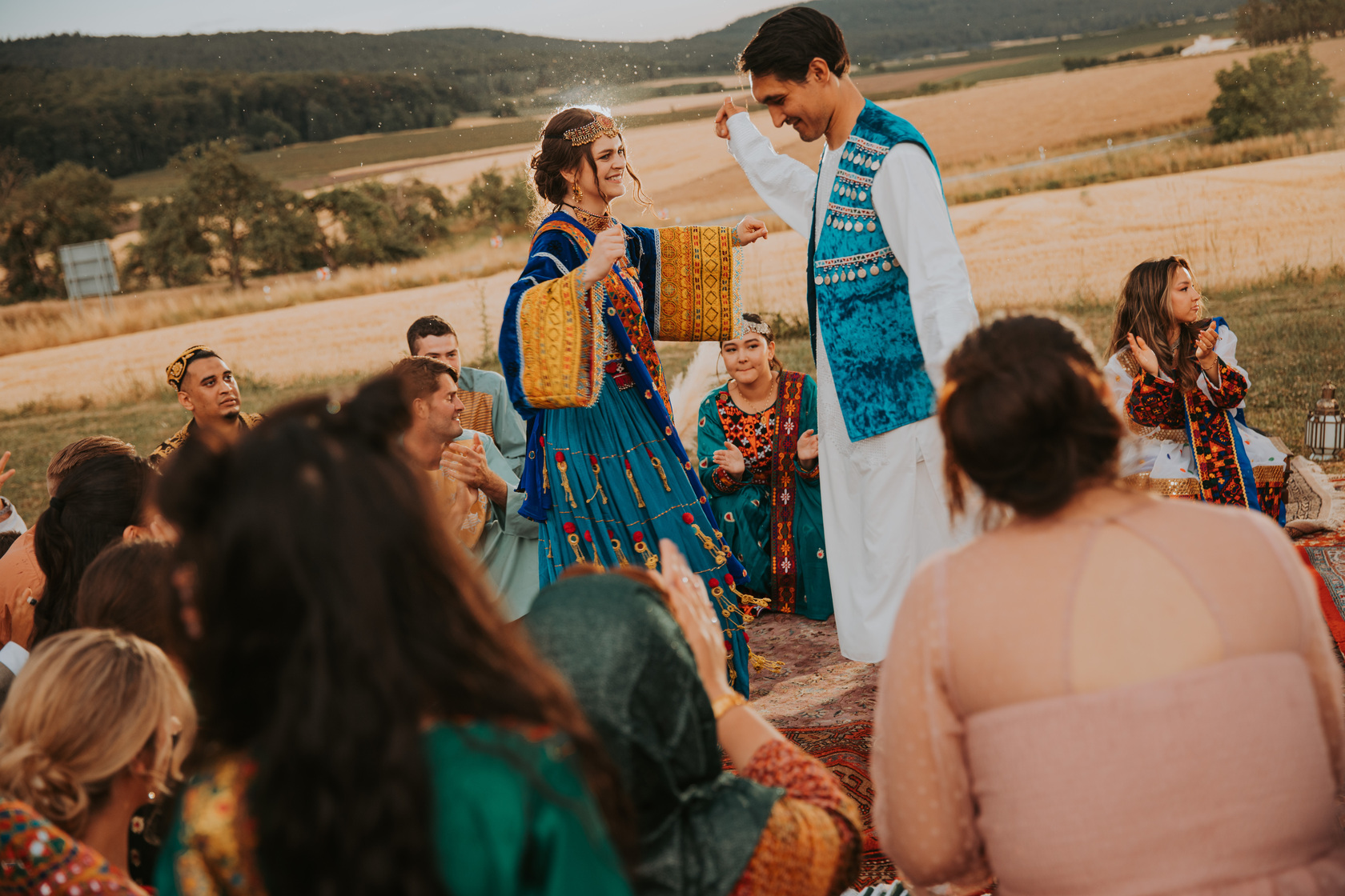 Ein Mann und eine Frau in traditioneller, farbenfroher Kleidung tanzen fröhlich bei einem Fest im Freien in der Nähe von Frankfurt. Sie sind von sitzenden Menschen umgeben, die klatschen und lächeln. Im Hintergrund ist eine malerische Landschaft mit Feldern und sanften Hügeln unter einem leicht bewölkten Himmel in der Abenddämmerung zu sehen. - Elis Wedding - Hochzeitsfotograf Frankfurt