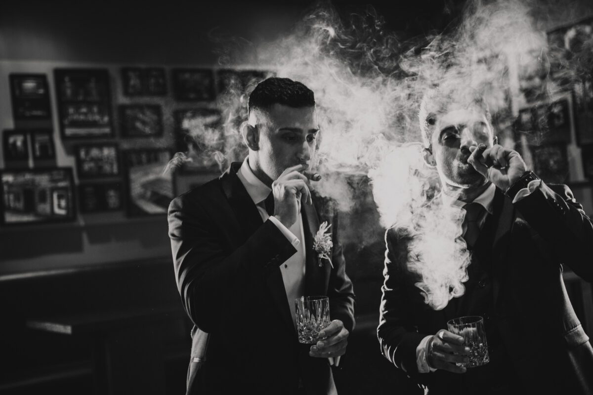Zwei Männer in Anzügen rauchen Zigarren und halten Whiskygläser in einem schwach beleuchteten Raum, wahrscheinlich Teil einer Fotoshooting-Session. Beide Männer atmen dichten Rauch aus, der ihre Gesichter teilweise verdeckt. Der Hintergrund ist verschwommen und zeigt gerahmte Bilder an einer Wand. Das Schwarzweißfoto erzeugt eine dramatische und klassische Atmosphäre. - Elis Wedding - Hochzeitsfotograf Frankfurt