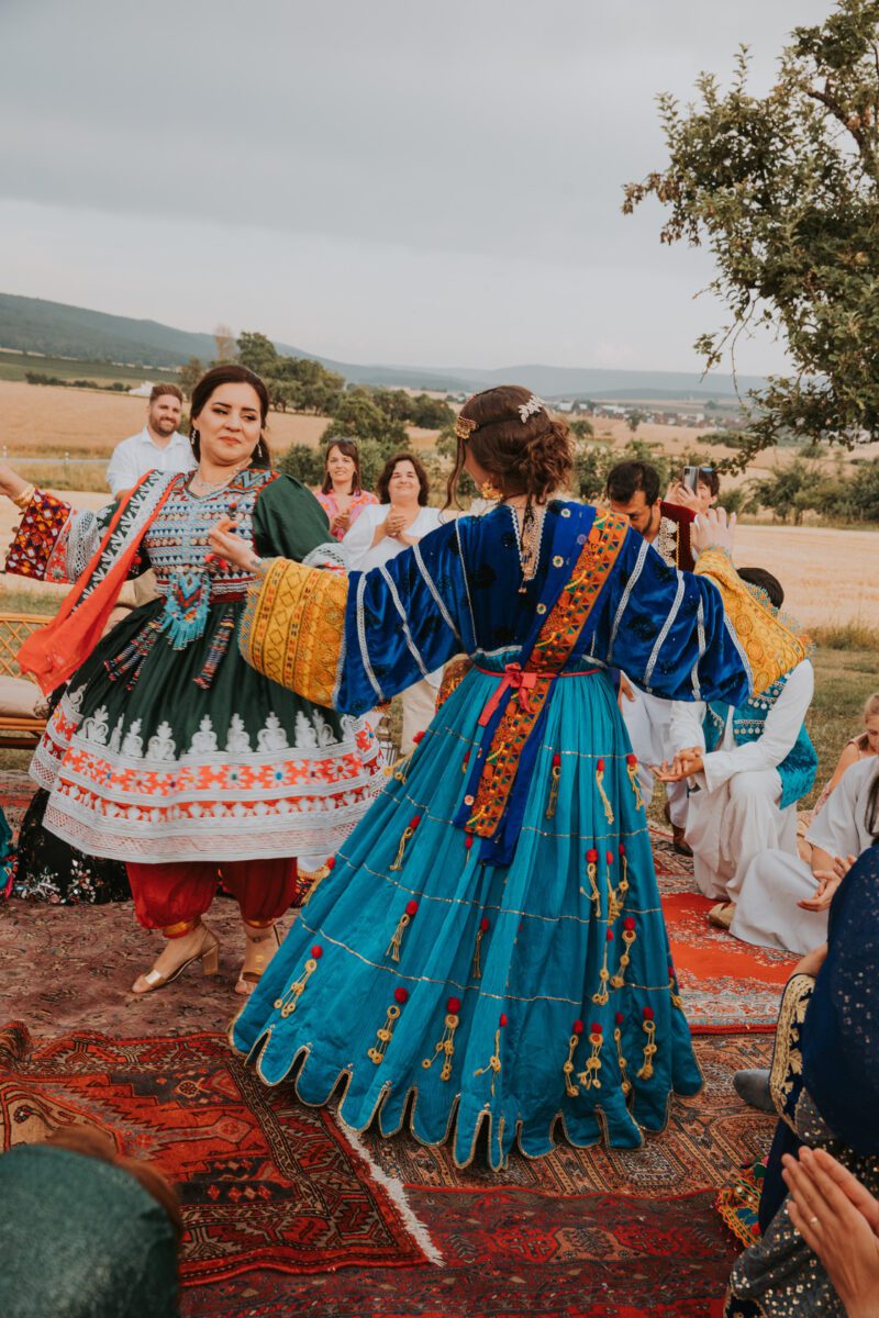 Zwei Frauen in farbenfroher traditioneller Kleidung tanzen fröhlich bei einem Treffen im Freien. Eine trägt ein grünes Kleid, die andere ein blaues Kleid mit gelben Ärmeln. Die Veranstaltung findet auf einem großen Teppich auf einem Feld in der Nähe von Frankfurt statt, die Zuschauer sitzen und stehen unter einem bewölkten Himmel. - Elis Wedding - Hochzeitsfotograf Frankfurt