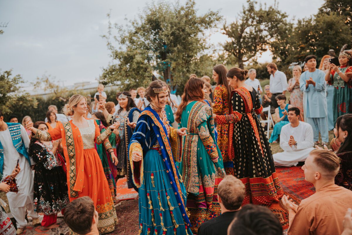 Eine Gruppe von Menschen in farbenfroher, traditioneller Kleidung nimmt an einer Feier im Freien teil, die an eine fröhliche standesamtliche Trauung erinnert. Mehrere Frauen tragen farbenfrohe Kleider, die mit komplizierten Mustern und Schmuck verziert sind, tanzen und lächeln. Männer und Kinder, die in verschiedene traditionelle Gewänder gekleidet sind, schauen zu und nehmen an den Festlichkeiten teil. Bäume und Grünflächen umgeben das Ereignis. - Elis Wedding - Hochzeitsfotograf Frankfurt