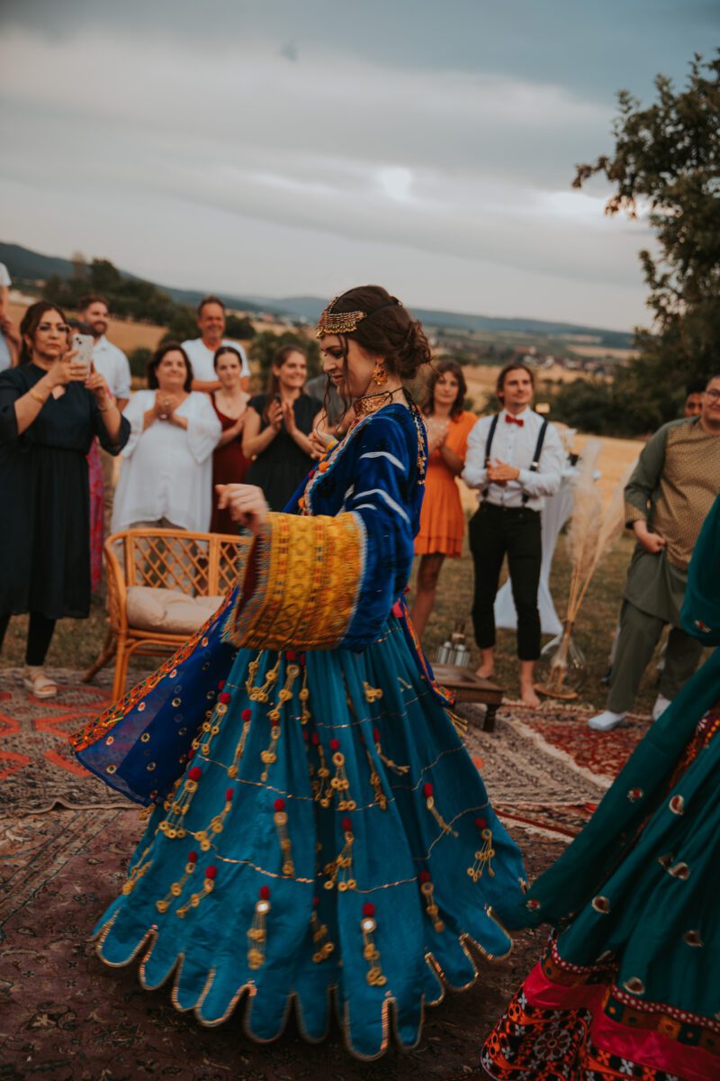 Eine Frau in farbenfroher, traditioneller Kleidung tanzt im Freien auf einem Teppich, umgeben von einer Gruppe klatschender und zuschauender Menschen. Die Kulisse ist eine hügelige, ländliche Gegend mit vereinzelten Bäumen und einem bewölkten Himmel. Männer und Frauen in legerer und traditioneller Kleidung genießen den Moment, perfekt für ein spontanes Fotoshooting. - Elis Wedding - Hochzeitsfotograf Frankfurt