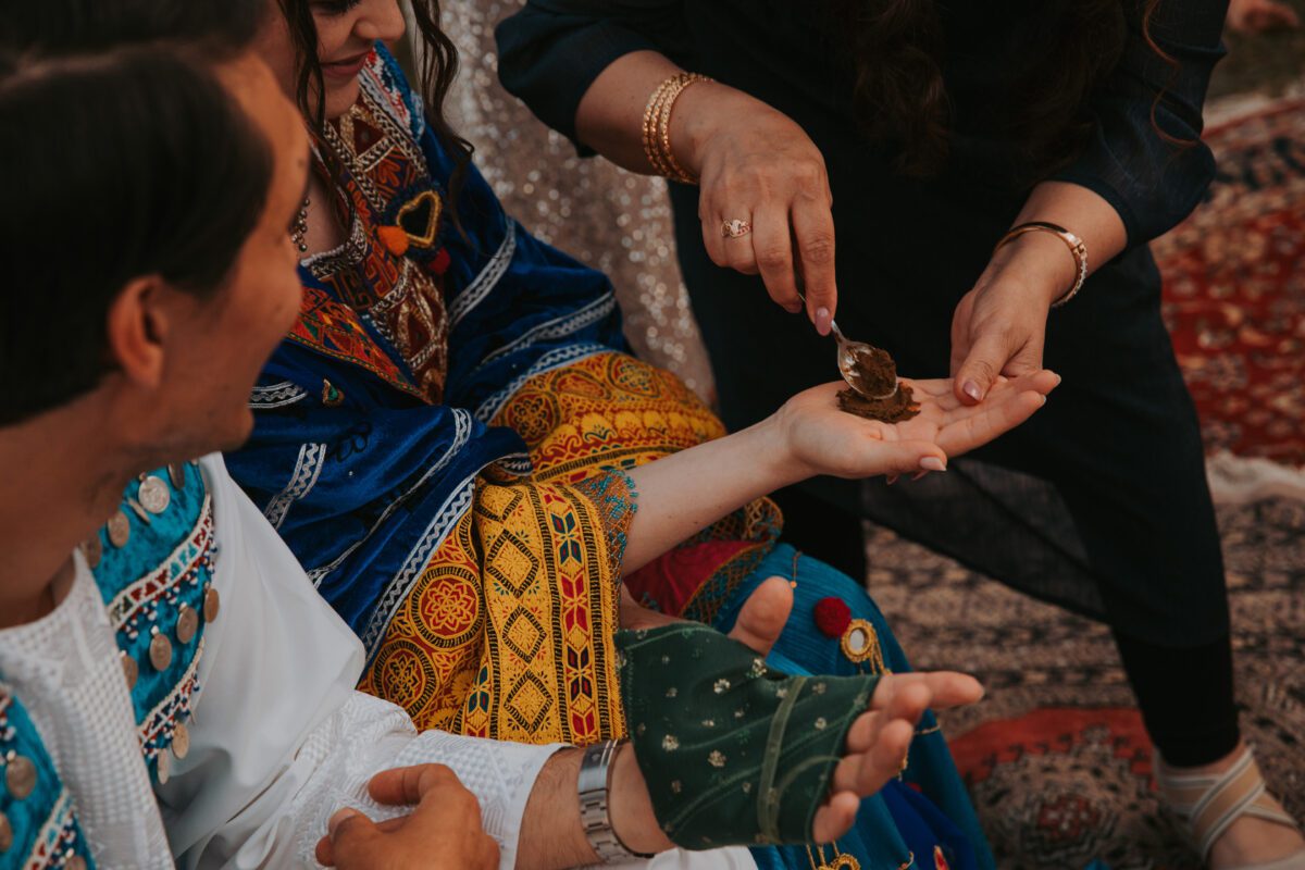 Zwei Menschen, in farbenfrohe traditionelle Gewänder gekleidet, nehmen an einer kulturellen Zeremonie teil. Die Hand einer Person wird von einer anderen Person mit einem Löffel mit einem Henna-Muster verziert. Die Szene ist intim und festlich und erinnert an ein Fotoshooting mit komplizierten Mustern sowohl auf der Kleidung als auch auf den Hintergrundtextilien. - Elis Wedding - Hochzeitsfotograf Frankfurt