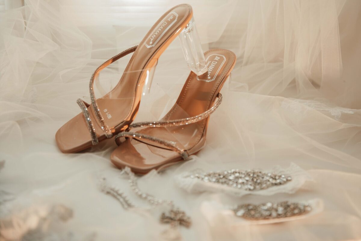 Ein Paar eleganter Riemchensandalen mit transparenten und metallischen Akzenten wird auf einem weichen, transparenten Stoffhintergrund präsentiert. Zarter Schmuck, darunter eine funkelnde Halskette und ein passendes Armband, liegt vor den Schuhen und unterstreicht das anspruchsvolle Ambiente, das zur Fotografie-Kollektion jedes Frankfurter Modebegeisterten passt. - Elis Wedding - Hochzeitsfotograf Frankfurt