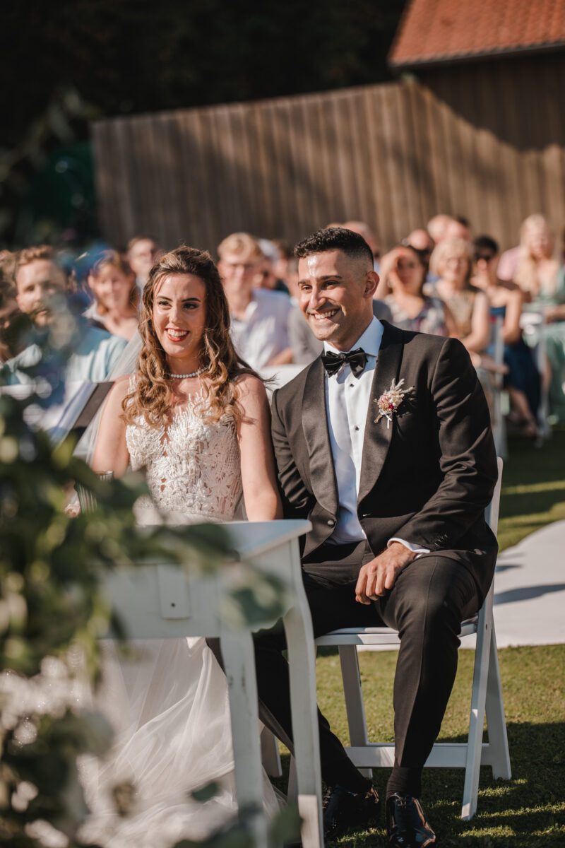 Braut und Bräutigam sitzen während einer standesamtlichen Trauung im Freien. Die Braut trägt ein weißes Kleid mit aufwendigen Spitzendetails und der Bräutigam einen schwarzen Anzug mit Fliege. Beide lächeln. Hinter ihnen sitzen Gäste in farbenfroher Kleidung unter einem sonnigen Himmel, im Hintergrund ist eine Holzkonstruktion zu sehen. - Elis Wedding - Hochzeitsfotograf Frankfurt