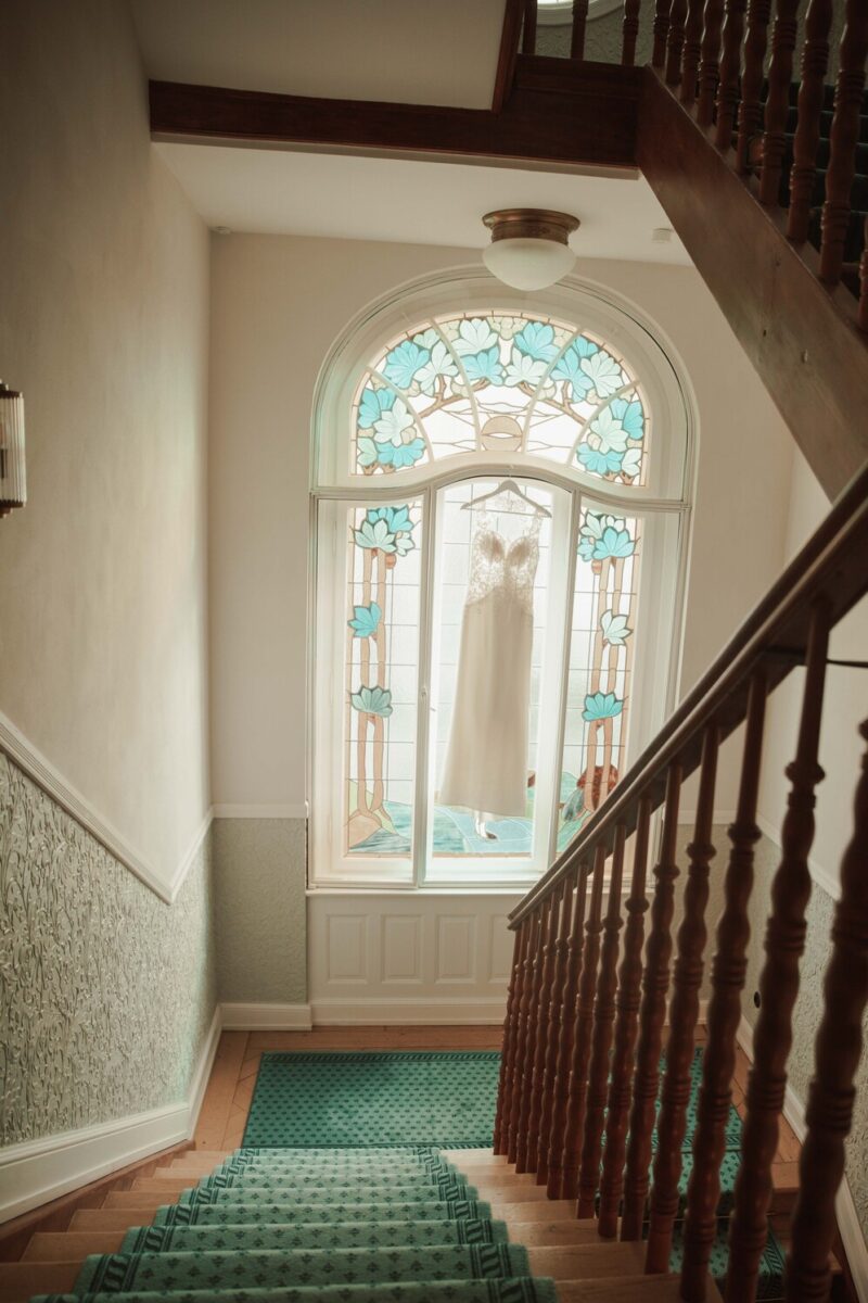 Ein Brautkleid hängt vor einem großen, kunstvoll verzierten Buntglasfenster mit floralen Mustern in Blau- und Weißtönen. Das Fenster befindet sich am Treppenabsatz einer Treppe mit Holzgeländer und grünem Teppich. Natürliches Licht erhellt den Bereich und macht ihn perfekt für Fotografien, die jedes kleinste Detail einfangen. - Elis Wedding - Hochzeitsfotograf Frankfurt