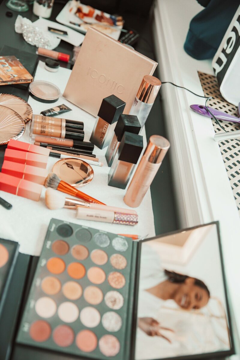 Ein Schminktisch, der mit verschiedenen Make-up-Produkten bedeckt ist, darunter Lidschattenpaletten, Foundation-Flaschen, Pinsel und Lippenstifte. Im Vordergrund ist eine Lidschattenpalette mit neutralen Tönen geöffnet, und ein Spiegel darauf reflektiert das lächelnde Gesicht einer Person, die sich vor ihrer standesamtlichen Trauung schminkt. - Elis Wedding - Hochzeitsfotograf Frankfurt