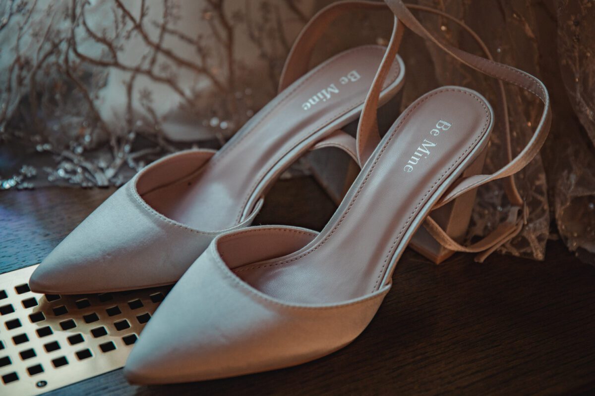 Ein Paar elegante, hellbeige High Heels mit spitzen Zehen und Riemchen stehen auf einem Holzboden. Auf den Innensohlen steht „Be Mine“. Im Hintergrund fällt ein Spitzenstoff mit Blumenmuster sanft herab und fängt einen perfekten Moment für ein Fotoshooting ein. - Elis Wedding - Hochzeitsfotograf Frankfurt
