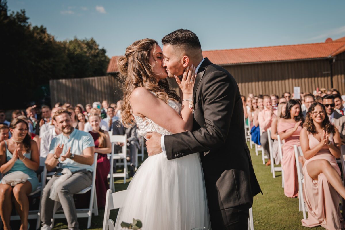 Braut und Bräutigam küssen sich während einer Hochzeitszeremonie im Freien, die von einem Hochzeitsfotografen festgehalten wurde. Die Braut in einem weißen Kleid und der Bräutigam in einem schwarzen Anzug stehen vor sitzenden Gästen, die klatschen und lächeln. Die sonnige Frankfurter Kulisse zeichnet sich durch einen klaren Himmel, eine Holzkonstruktion und Bäume im Hintergrund aus. - Elis Wedding - Hochzeitsfotograf Frankfurt