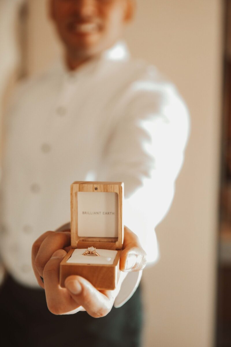 Eine Person in einem weißen Button-Down-Hemd hält eine offene Ringschachtel aus Holz in die Kamera. Die Schachtel, die sich perfekt zum Fotografieren eignet, enthält einen Diamantring und auf der Innenseite ist „BRILLIANT EARTH“ aufgedruckt. Der sanft beleuchtete Hintergrund schafft eine warme und intime Atmosphäre. - Elis Wedding - Hochzeitsfotograf Frankfurt