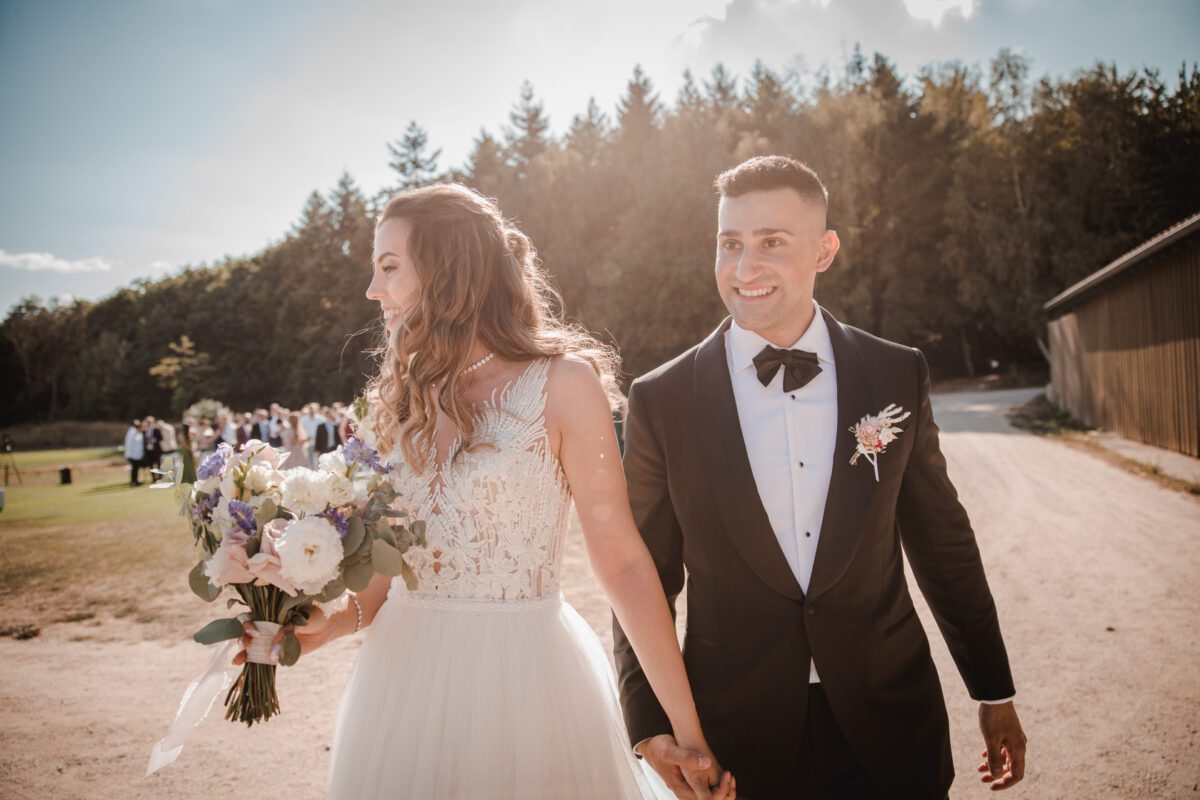 Braut und Bräutigam halten sich an den Händen und lächeln auf einem sonnenbeschienenen Weg bei einer Hochzeit im Freien. Die Braut trägt ein weißes Kleid mit Spitzendetails und hält einen Blumenstrauß, während der Bräutigam einen schwarzen Smoking mit Fliege und Ansteckblume trägt. Im Hintergrund sind Gäste zu sehen, umgeben von Bäumen und Grün, die diesen wunderschönen Moment des Kennenlernens durch Fotografie in Frankfurt festhalten. - Elis Wedding - Hochzeitsfotograf Frankfurt