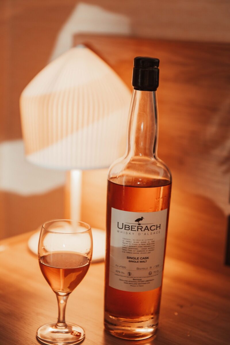 Eine Flasche Uberach Whiskey d'Alsace Single Malt steht auf einer Holzoberfläche neben einem gefüllten Whiskeyglas. Im Hintergrund steht eine sanft beleuchtete Tischlampe mit weißem Schirm, die einen warmen Schein über die Szene wirft. Diese gemütliche Umgebung mit ihren durchgehend warmen Farbtönen ist perfekt für eine intime Hochzeitsfotografen-Session. - Elis Wedding - Hochzeitsfotograf Frankfurt