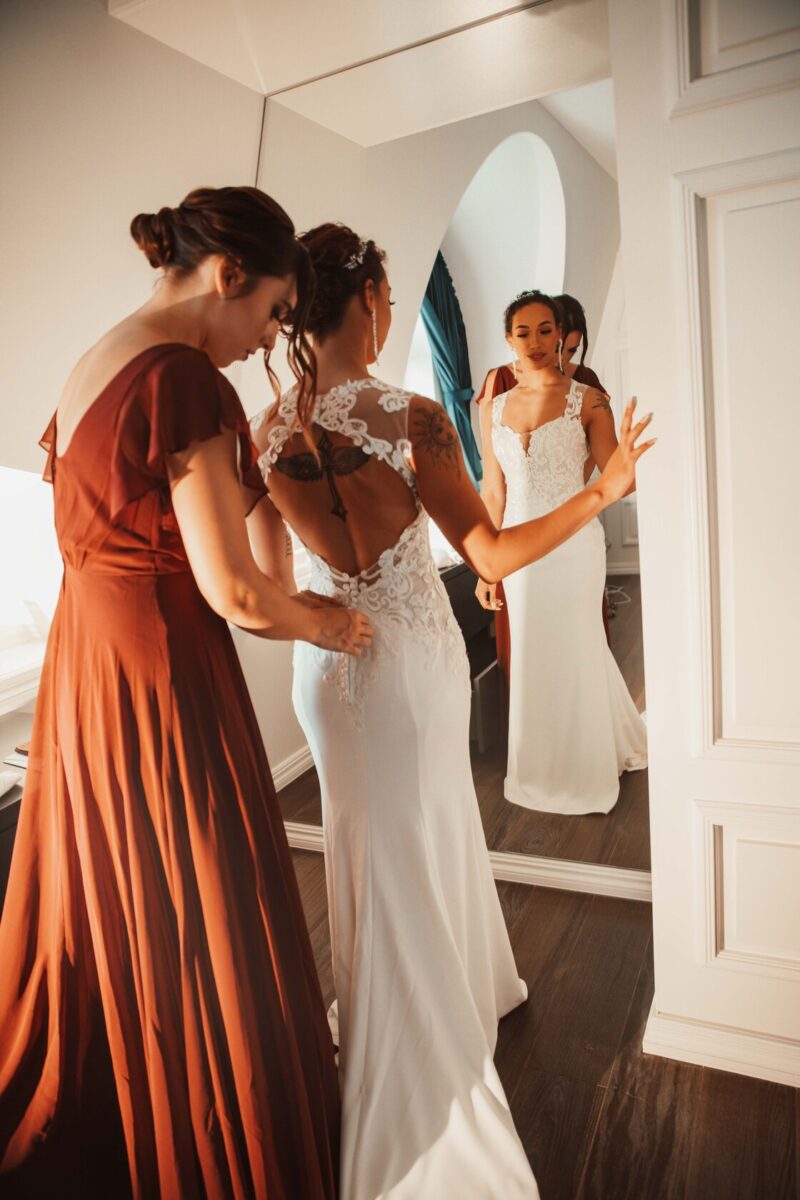 Eine Braut in einem weißen Kleid mit Spitzendetails steht vor einem Spiegel und bereitet sich auf ihre standesamtliche Trauung vor. Eine Frau in einem langen, braunen Kleid hilft mit der Rückseite ihres Kleides. Der Raum hat weiße Wände und einen Holzboden, was eine warme Atmosphäre schafft. Die Braut betrachtet ihr Spiegelbild mit einem ruhigen Gesichtsausdruck, bereit für den Hochzeitsfotografen, um dies festzuhalten - Elis Wedding - Hochzeitsfotograf Frankfurt
