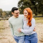 Ein Paar geht Arm in Arm durch eine grasbewachsene Außenanlage, als ob es gerade seine standesamtliche Trauung hinter sich hätte. Der Mann, der eine Brille und ein hellgrünes Hemd trägt, sieht die Frau lächelnd an. Die Frau, mit langen roten Haaren und einem hellblauen Hemd, lächelt herzlich. Im Hintergrund sind Bäume und ein Holzzaun zu sehen. - Elis Wedding - Hochzeitsfotograf Frankfurt