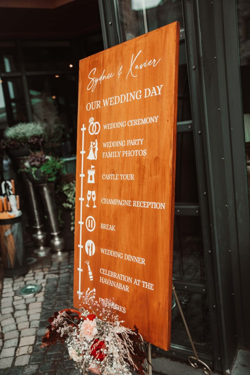 An einer Glaswand hängt ein hölzernes Schild mit dem Hochzeitsplan. Das Schild mit der Überschrift „Unser Hochzeitstag“ listet die Ereignisse auf, darunter die Hochzeitszeremonie, Hochzeitsfotograf, Familienfotos, Schlossführung, Champagnerempfang, Pause, Hochzeitsessen, Feier in der Havanabar und Feuerwerk. Blumenarrangements liegen am Sockel. - Elis Wedding - Hochzeitsfotograf Frankfurt
