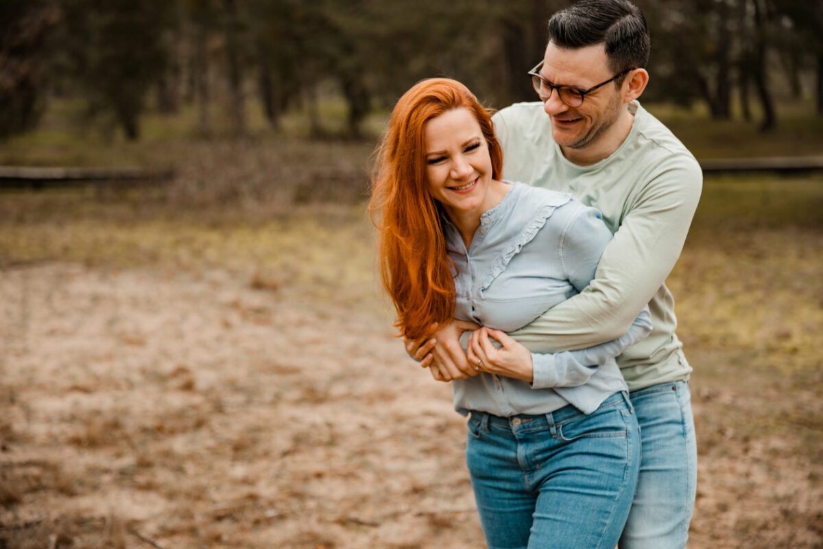 Ein Paar steht im Freien mit Bäumen im Hintergrund. Die Frau mit langen roten Haaren, einem hellblauen Top und Jeans lächelt, während der Mann mit kurzen dunklen Haaren und Brille, einem hellgrünen Shirt und Jeans sie von hinten umarmt. Beide wirken glücklich und genießen den Moment während ihres Fotoshootings in Frankfurt. - Elis Wedding - Hochzeitsfotograf Frankfurt