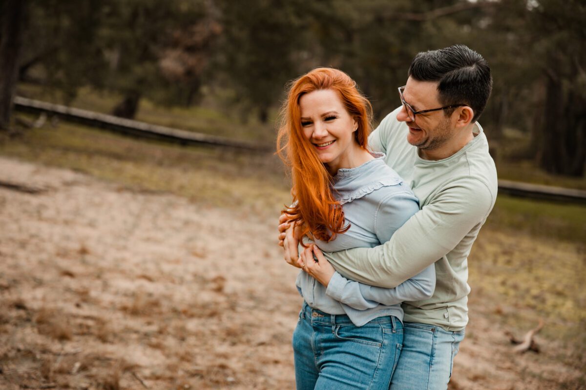 Ein Paar lächelt und umarmt sich im Freien während eines Fotoshootings. Die Frau mit den langen roten Haaren trägt eine hellblaue Bluse und hellblaue Jeans, während der Mann mit den kurzen dunklen Haaren und der Brille ein hellgrünes Hemd und blaue Jeans trägt. Sie stehen auf sandigem, grasbewachsenem Gelände mit Bäumen im Hintergrund. - Elis Wedding - Hochzeitsfotograf Frankfurt