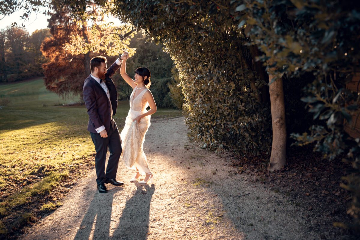 Ein Paar tanzt während des Fotoshootings anmutig auf einem sonnenbeschienenen, schattigen Weg, umgeben von üppigem Grün. Die Frau in einem weißen Kleid wirbelt unter der Führung des Mannes herum. Der Mann, der für ihre standesamtliche Trauung einen dunklen Anzug trägt, lächelt, während er führt. Warmes Sonnenlicht fällt durch die Bäume und wirft einen malerischen goldenen Schimmer auf den Weg und das Paar. - Elis Wedding - Hochzeitsfotograf Frankfurt