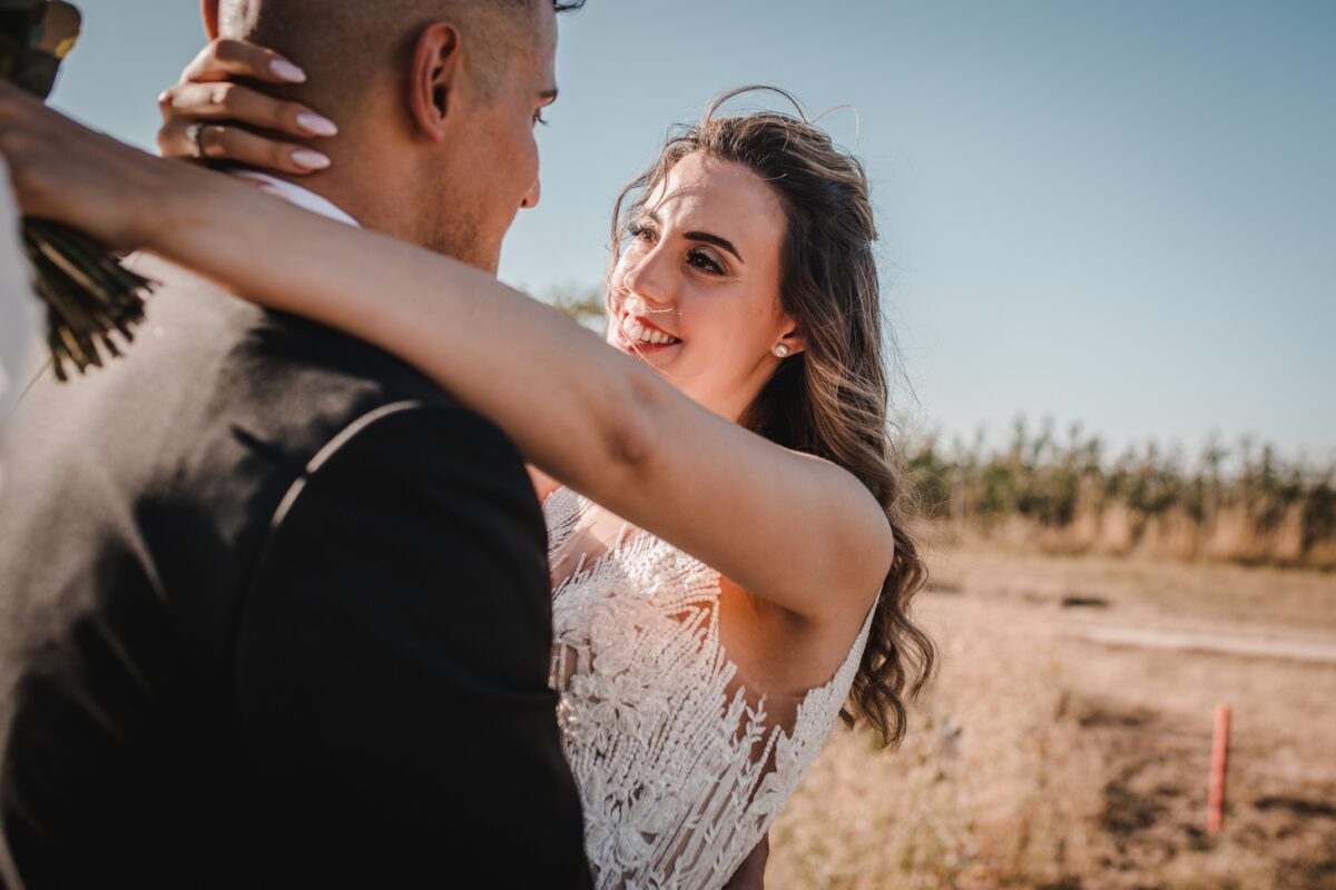 Braut und Bräutigam teilen einen intimen Moment im Freien an einem sonnigen Tag, perfekt festgehalten von ihrem Hochzeitsfotografen. Die Braut, in einem weißen Spitzenkleid, blickt den Bräutigam mit einem freudigen Lächeln an, die Arme um seine Schultern gelegt. Der Bräutigam, in einem schwarzen Anzug, blickt ihr vor einem klaren blauen Himmel und grasbedeckten Feldern entgegen. - Elis Wedding - Hochzeitsfotograf Frankfurt