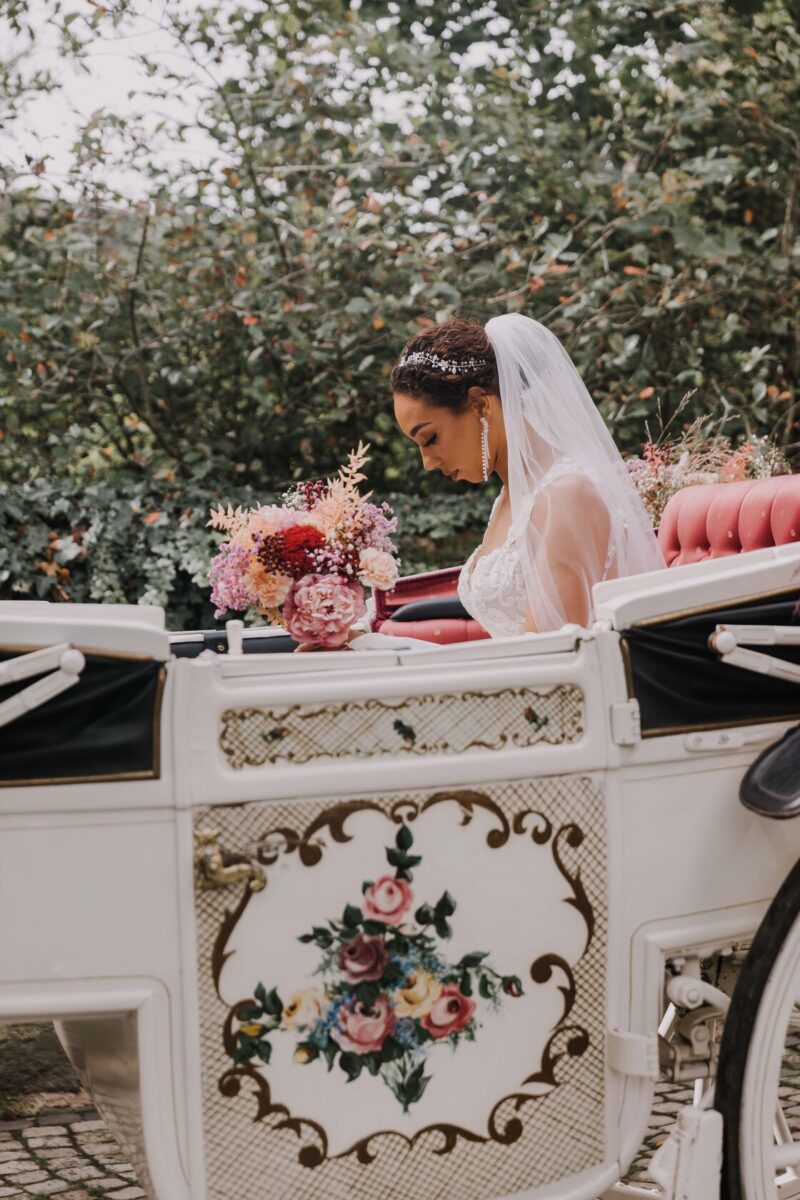 Eine Braut in einem weißen Spitzenhochzeitskleid und Schleier sitzt auf einem roten gepolsterten Sitz in einer verzierten weißen Kutsche, die mit Blumenmustern verziert ist. Sie hält einen Strauß rosa und roter Blumen in der Hand und blickt während ihrer standesamtlichen Trauung nachdenklich nach unten. Im Hintergrund ist grünes Laub zu sehen. - Elis Wedding - Hochzeitsfotograf Frankfurt
