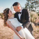 Ein Paar in Hochzeitskleidung posiert an einem sonnigen Tag im Freien. Die Braut in einem weißen Spitzenkleid wird vom Bräutigam, der einen schwarzen Smoking und eine Fliege trägt, umarmt. Sie schauen sich lächelnd in die Augen, umgeben von Grün und einem klaren blauen Himmel – ein perfekter Moment, eingefangen von ihrem Hochzeitsfotografen in Frankfurt. - Elis Wedding - Hochzeitsfotograf Frankfurt