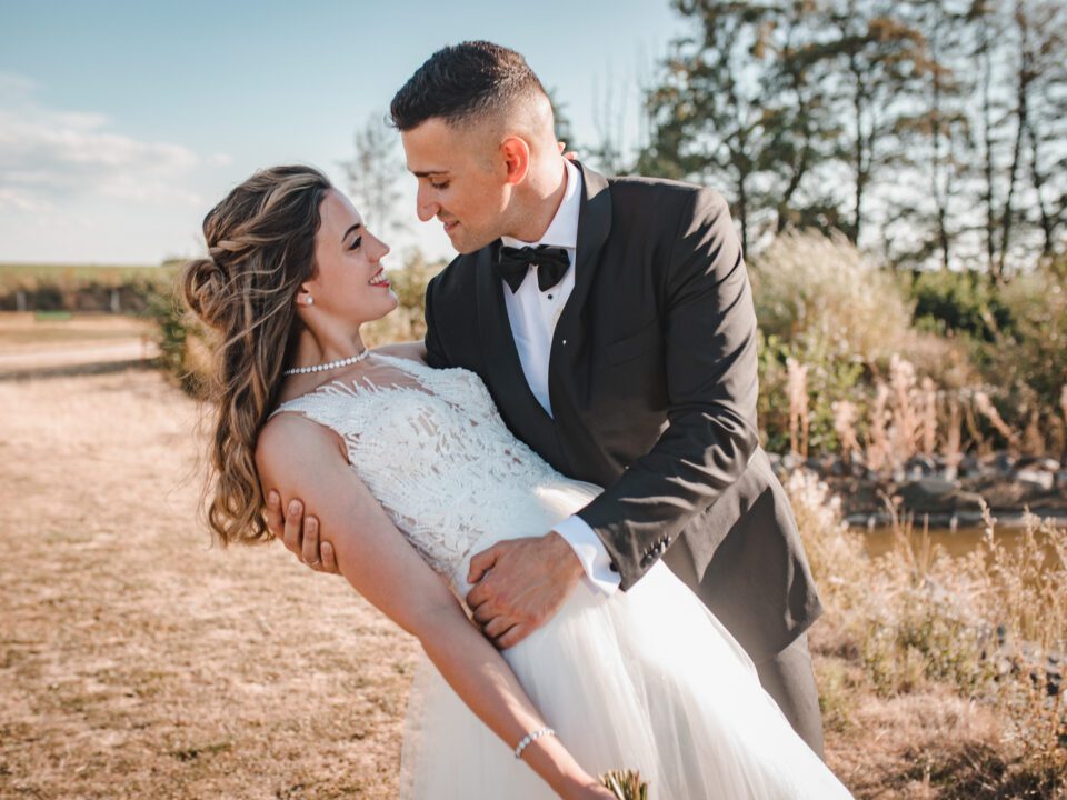 Ein Paar in Hochzeitskleidung posiert an einem sonnigen Tag im Freien. Die Braut in einem weißen Spitzenkleid wird vom Bräutigam, der einen schwarzen Smoking und eine Fliege trägt, umarmt. Sie schauen sich lächelnd in die Augen, umgeben von Grün und einem klaren blauen Himmel – ein perfekter Moment, eingefangen von ihrem Hochzeitsfotografen in Frankfurt. - Elis Wedding - Hochzeitsfotograf Frankfurt