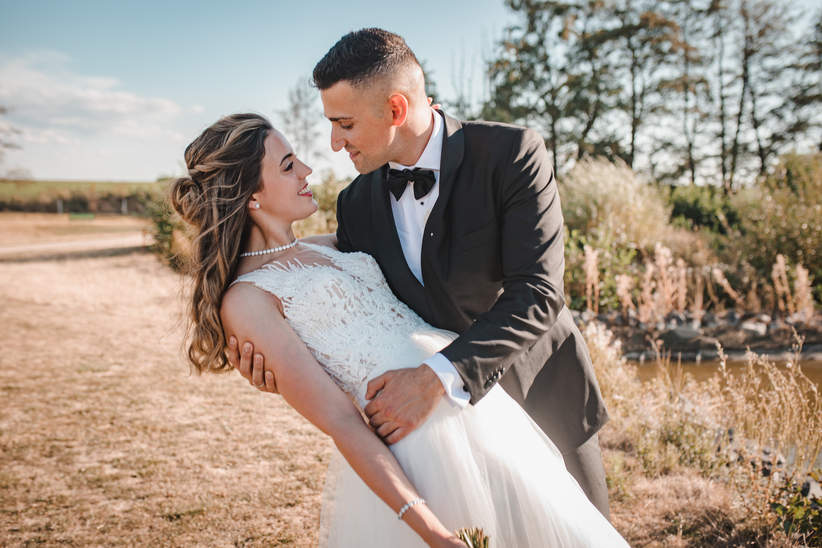 Ein Paar in Hochzeitskleidung posiert an einem sonnigen Tag im Freien. Die Braut in einem weißen Spitzenkleid wird vom Bräutigam, der einen schwarzen Smoking und eine Fliege trägt, umarmt. Sie schauen sich lächelnd in die Augen, umgeben von Grün und einem klaren blauen Himmel – ein perfekter Moment, eingefangen von ihrem Hochzeitsfotografen in Frankfurt. - Elis Wedding - Hochzeitsfotograf Frankfurt