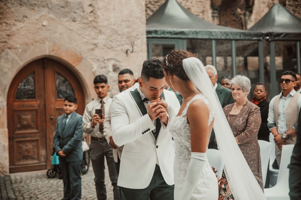 Ein Hochzeitsfotograf fängt den Moment ein, in dem ein Bräutigam im weißen Smoking seiner Braut im weißen Spitzenkleid während ihrer Trauung im Freien die Hand küsst. Die Braut, geschmückt mit einem langen Schleier und Handschuhen, steht zwischen Gästen, darunter eine ältere Frau und andere in Abendgarderobe. Im Hintergrund sind eine gewölbte Holztür und Steinmauern zu sehen. - Elis Wedding - Hochzeitsfotograf Frankfurt