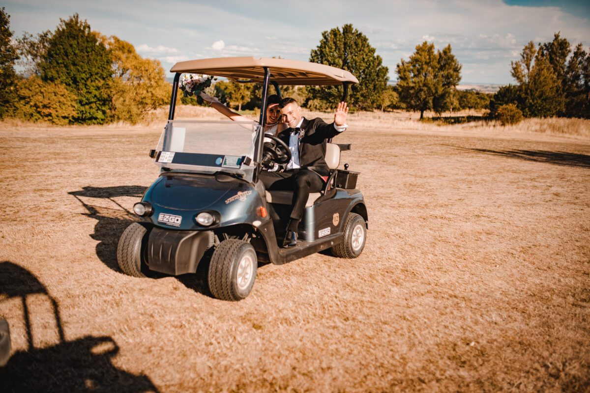 Ein Paar in formeller Kleidung winkt freudig von einem schwarzen Golfwagen auf einer trockenen, grasbedeckten Wiese. Die Frau, die ein weißes Kleid und einen Schleier trägt, hält einen Blumenstrauß, während der Mann in einem dunklen Anzug und einer Fliege fährt. Auf einer wunderschönen Fotografie in der Nähe von Frankfurt während ihrer standesamtlichen Trauung sind sie umgeben von Bäumen mit Herbstlaub unter einem klaren Himmel. - Elis Wedding - Hochzeitsfotograf Frankfurt
