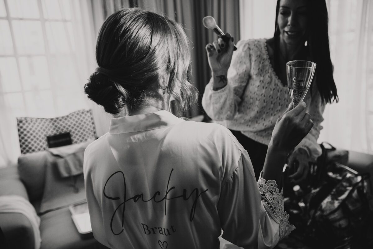 Eine Braut mit elegant hochgesteckten Haaren, trägt ein Gewand mit der Aufschrift „Jacky Braut“ auf dem Rücken, hält ein Champagnerglas und steht einer Frau mit langen Haaren gegenüber, die sich schminkt. In dem Raum mit großen Fenstern und Vorhängen ruhen verschiedene Gegenstände und Taschen auf einem Sofa dahinter. Der Hochzeitsfotograf fängt jeden Moment dieses intimen Auftakts zu ihrem Stand ein - Elis Wedding - Hochzeitsfotograf Frankfurt