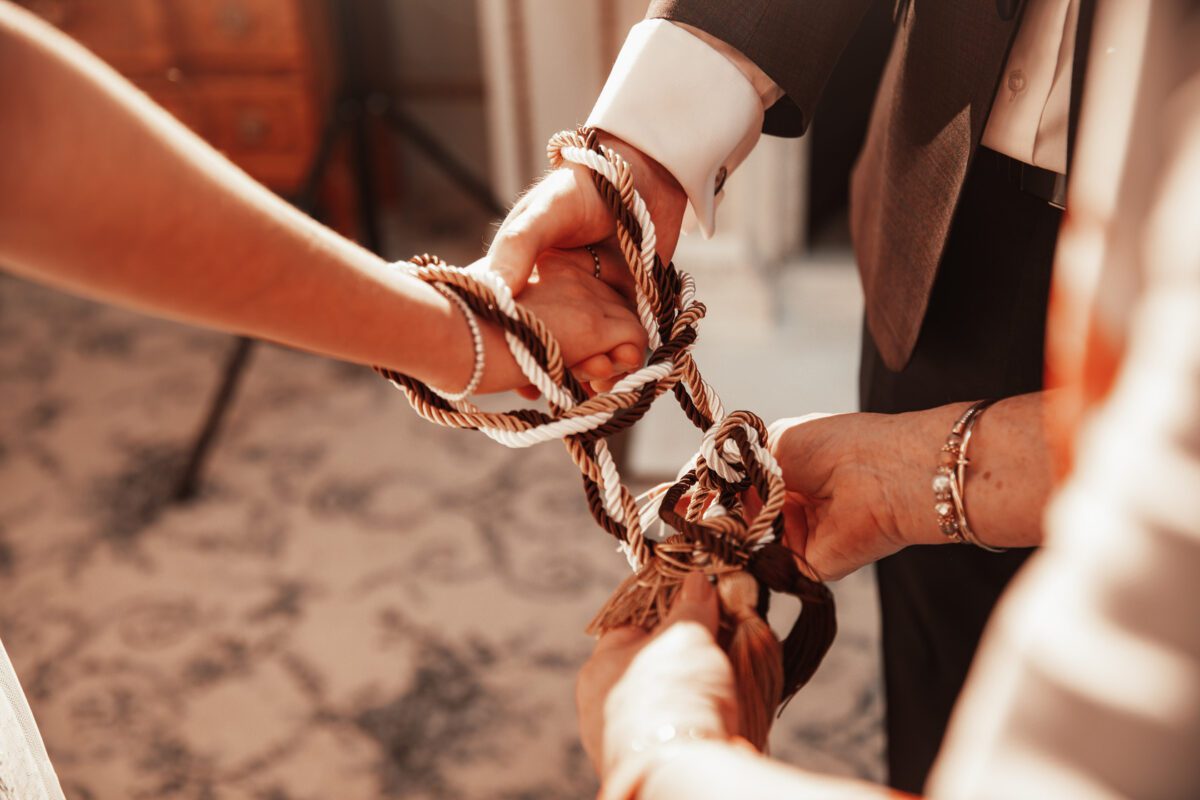 Nahaufnahme der Hände von drei Personen, von denen zwei ein Seil halten, das kunstvoll um die Hände der dritten Person gebunden ist, was eine Einheits- oder Handfasting-Zeremonie symbolisiert. Der sanft beleuchtete Hintergrund besteht aus gemustertem Teppich und Holzmöbeln. Diese atemberaubende Fotografie fängt einen intimen Moment ein und ist perfekt für das Portfolio jedes Hochzeitsfotografen. - Elis Wedding - Hochzeitsfotograf Frankfurt