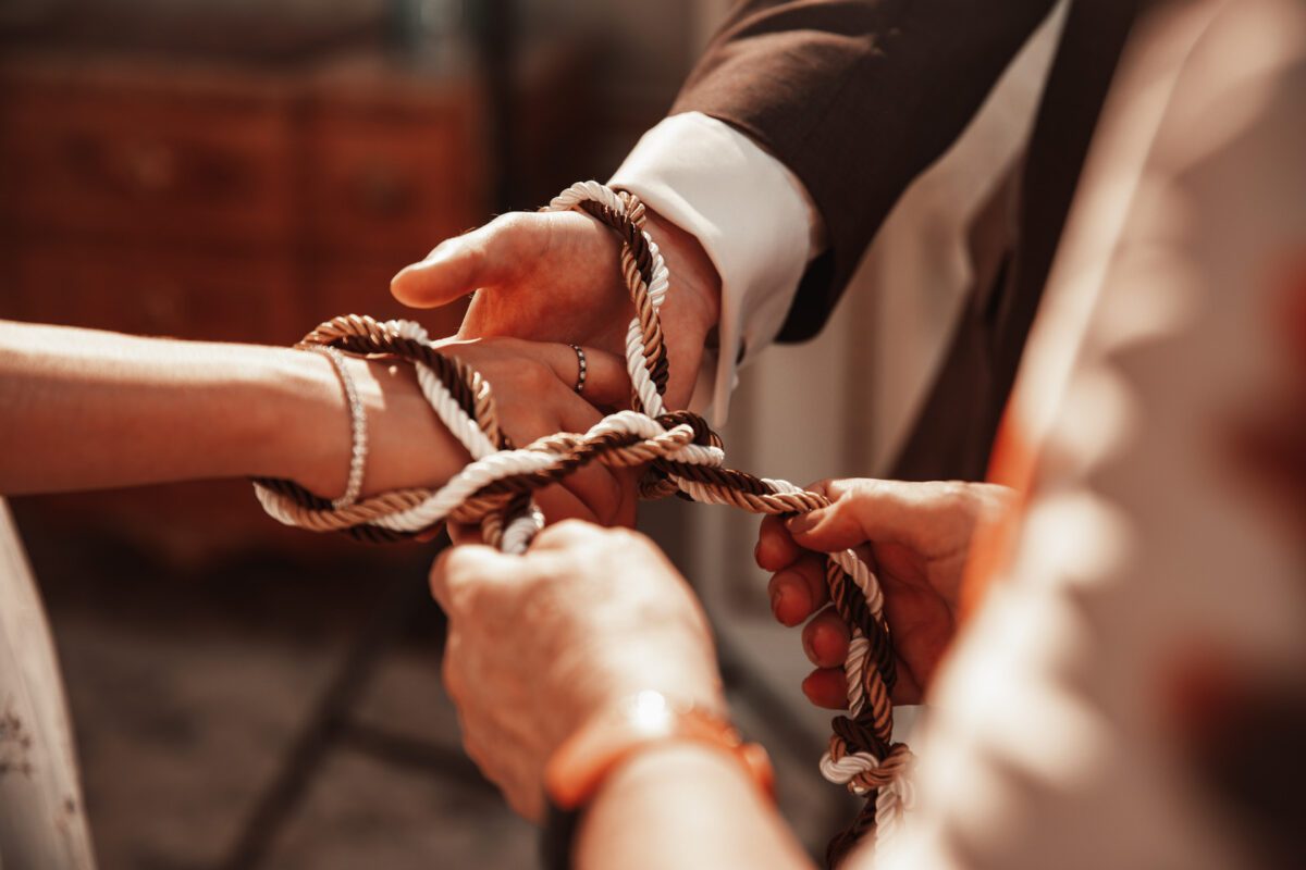 Nahaufnahme von vier Händen, die an einer Einheitszeremonie teilnehmen, wahrscheinlich einer standesamtlichen Trauung, bei der eine dicke, gedrehte Kordel um ihre Handgelenke gebunden wird. Die Kordel symbolisiert Einheit und Engagement. Aufgenommen von einem Hochzeitsfotografen in Frankfurt, verschwimmen im Hintergrund sanft die Holzmöbel und das warme Licht. - Elis Wedding - Hochzeitsfotograf Frankfurt