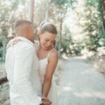 Ein Paar umarmt sich auf einem sonnenbeschienenen Waldweg. Die Frau im weißen Kleid lächelt und blickt nach unten, während sie die Hand des Mannes hält. Der Mann, von hinten zu sehen, trägt in einem weißen Hemd zum Charme des Augenblicks bei. Sonnenlicht fällt durch die grünen, belaubten Bäume und schafft eine ruhige und romantische Atmosphäre, perfekt für ein spontanes Fotoshooting in der Nähe von Frankfurt. - Elis Wedding - Hochzeitsfotograf Frankfurt