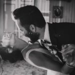 Ein Schwarzweißfoto eines tanzenden Paares. Der Mann im Anzug taucht die Frau in ein Abendkleid. Sie lehnt sich mit geschlossenen Augen und freudigem Gesichtsausdruck zurück. Sie halten sich an den Händen, und im Hintergrund sind gerahmte Bilder und kunstvolle Dekorationen zu sehen, die an eine elegante Kulisse erinnern, die von einem Hochzeitsfotografen aufgenommen wurde. - Elis Wedding - Hochzeitsfotograf Frankfurt