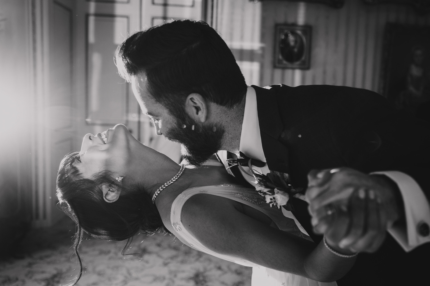 Ein Schwarzweißfoto eines tanzenden Paares. Der Mann im Anzug taucht die Frau in ein Abendkleid. Sie lehnt sich mit geschlossenen Augen und freudigem Gesichtsausdruck zurück. Sie halten sich an den Händen, und im Hintergrund sind gerahmte Bilder und kunstvolle Dekorationen zu sehen, die an eine elegante Kulisse erinnern, die von einem Hochzeitsfotografen aufgenommen wurde. - Elis Wedding - Hochzeitsfotograf Frankfurt