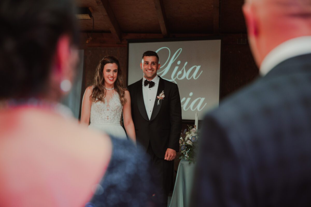 Eine Braut im weißen Kleid und ein Bräutigam im schwarzen Smoking stehen beide lächelnd zusammen und blicken in die Kamera. Im Hintergrund ist ein Bildschirm mit „Lisa“ und einem Teil eines anderen Namens zu sehen. Der Fokus liegt auf dem Paar, wobei zwei Personen im Vordergrund unscharf sind und ihnen möglicherweise bei ihrer standesamtlichen Trauung zuschauen. - Elis Wedding - Hochzeitsfotograf Frankfurt