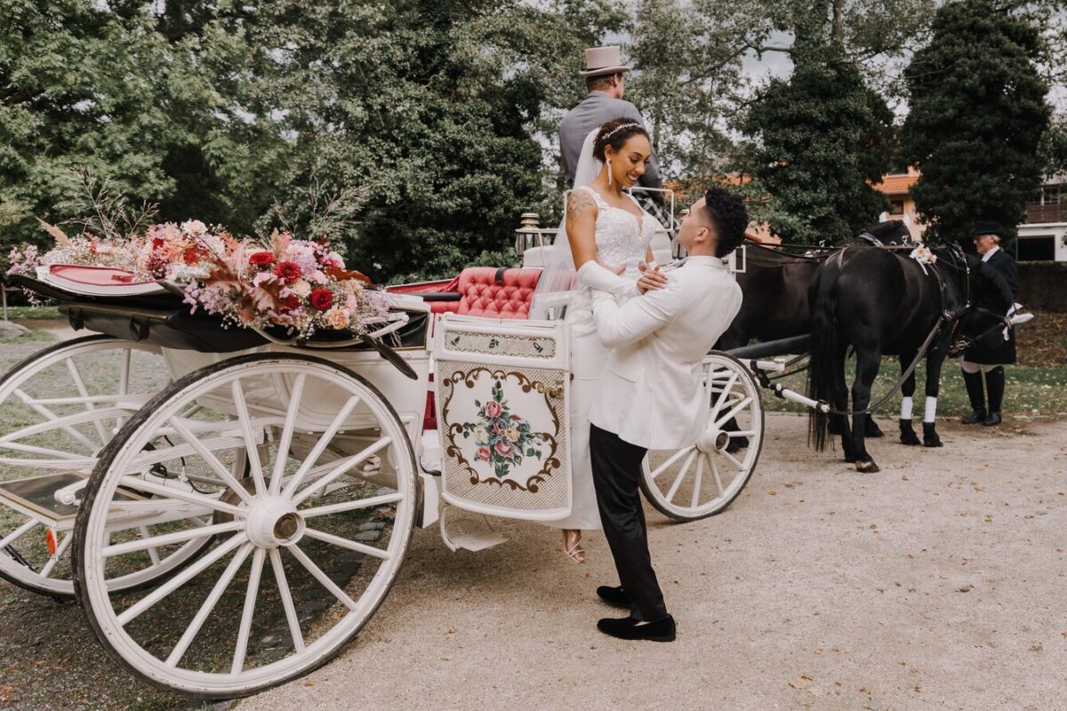Ein Bräutigam hebt seine Braut vor eine weiße, reich verzierte Kutsche, die mit rosa und roten Blumenarrangements geschmückt ist. Er trägt einen weißen Anzug und sie ein ärmelloses Hochzeitskleid mit langem Schleier. Die Kutsche wird von zwei schwarzen Pferden gezogen und der Kutscher hält die Zügel. Es ist ein perfekter Moment für den Hochzeitsfotografen, um die Kutsche zwischen Bäumen und Gebäuden im Hintergrund festzuhalten. - Elis Wedding - Hochzeitsfotograf Frankfurt