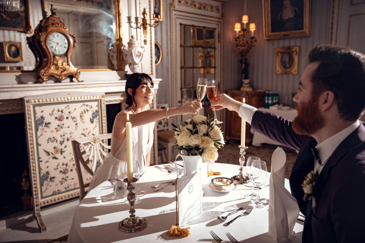 Braut und Bräutigam sitzen an einem elegant gedeckten Esstisch und stoßen mit Champagnergläsern an. Der Raum, der an das klassische Frankfurter Dekor erinnert, verfügt über eine kunstvolle Uhr und einen großen Strauß weißer Rosen. Kerzenlicht sorgt für einen warmen Schein und hebt den detailreichen Kaminsims in dieser exquisiten Fotografie dieses Hochzeitsfotografen hervor. - Elis Wedding - Hochzeitsfotograf Frankfurt