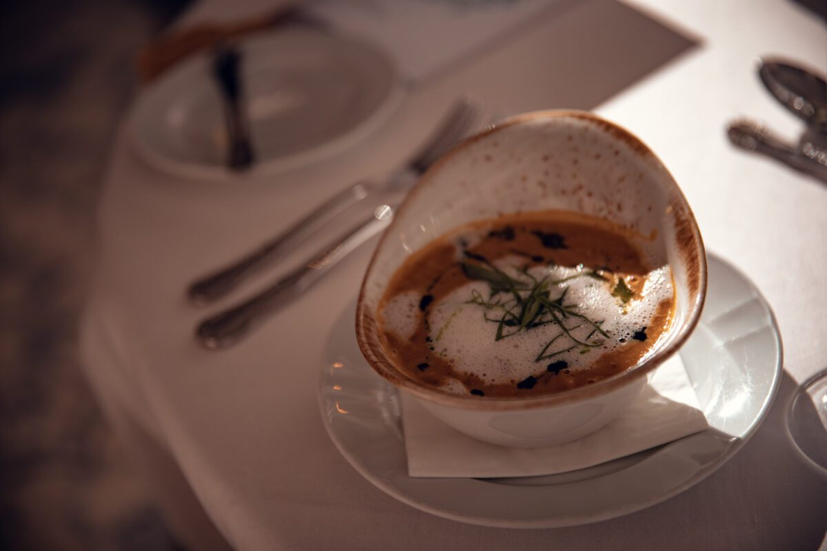Eine Nahaufnahme einer elegant präsentierten Schüssel cremiger Suppe, garniert mit grünen Kräutern und einer weißen Schaumkrone, auf einem weißen Teller mit einer Serviette darunter. Der Hintergrund, der an die Kunstfertigkeit eines Hochzeitsfotografen bei der Aufnahme gehobener Speisesäle erinnert, zeigt verschwommenes Geschirr. - Elis Wedding - Hochzeitsfotograf Frankfurt