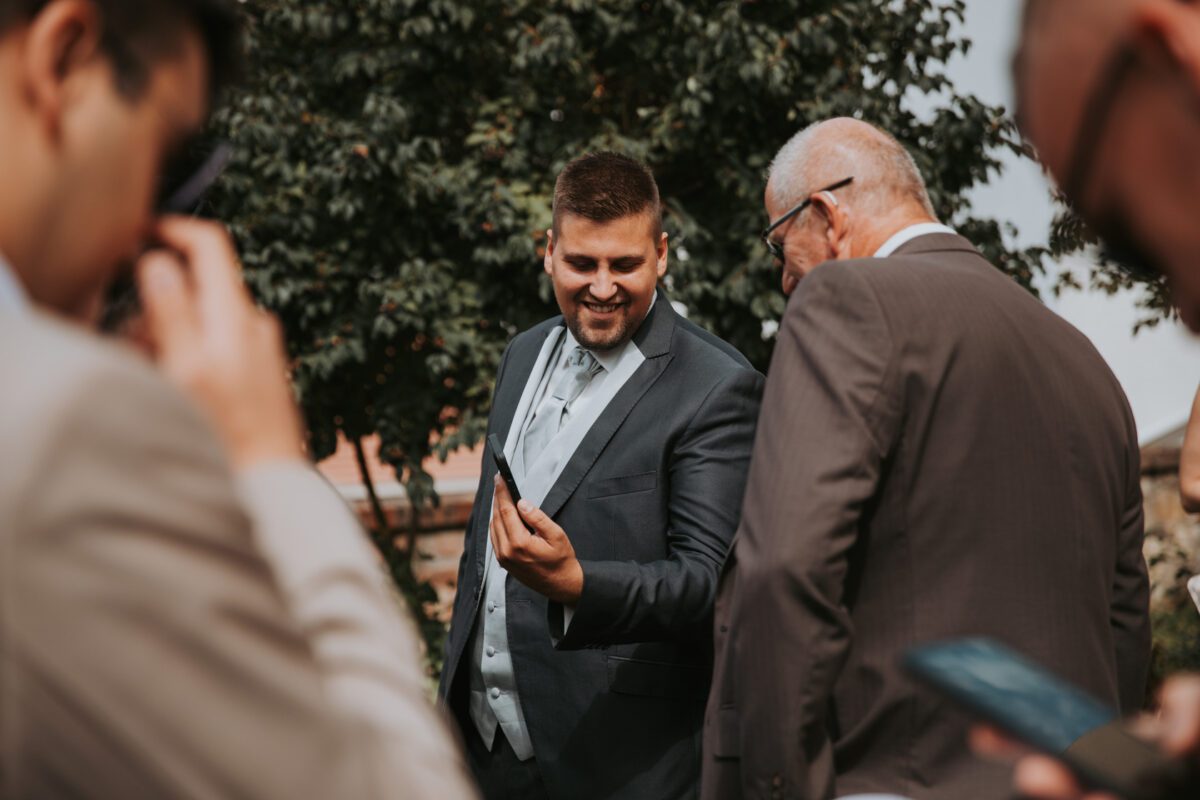 Ein Mann in einem grauen Anzug, der ein Telefon hält und lächelt, zeigt einem älteren Mann in einem braunen Anzug und mit Brille etwas. Sie sind im Freien, mit Bäumen im Hintergrund. Andere Personen, die teilweise im Vordergrund zu sehen sind, sind ebenfalls in formeller Kleidung gekleidet, wahrscheinlich bei einem Fotoshooting oder einer standesamtlichen Trauung. - Elis Wedding - Hochzeitsfotograf Frankfurt