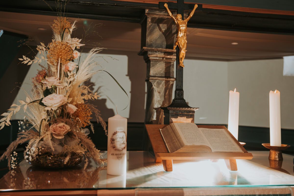 Ein Kirchenaltar, geschmückt mit einem Blumenarrangement, einem Kruzifix, zwei brennenden Kerzen und einer offenen Bibel auf einem Holzständer. Zu den Blumen gehören weiße Rosen, gelbe Blüten und Weizenhalme. Perfekt vom Hochzeitsfotografen eingefangen, strahlen die Kerzen ein sanftes Licht aus, während das Kruzifix fest hinter der Bibel steht. - Elis Wedding - Hochzeitsfotograf Frankfurt