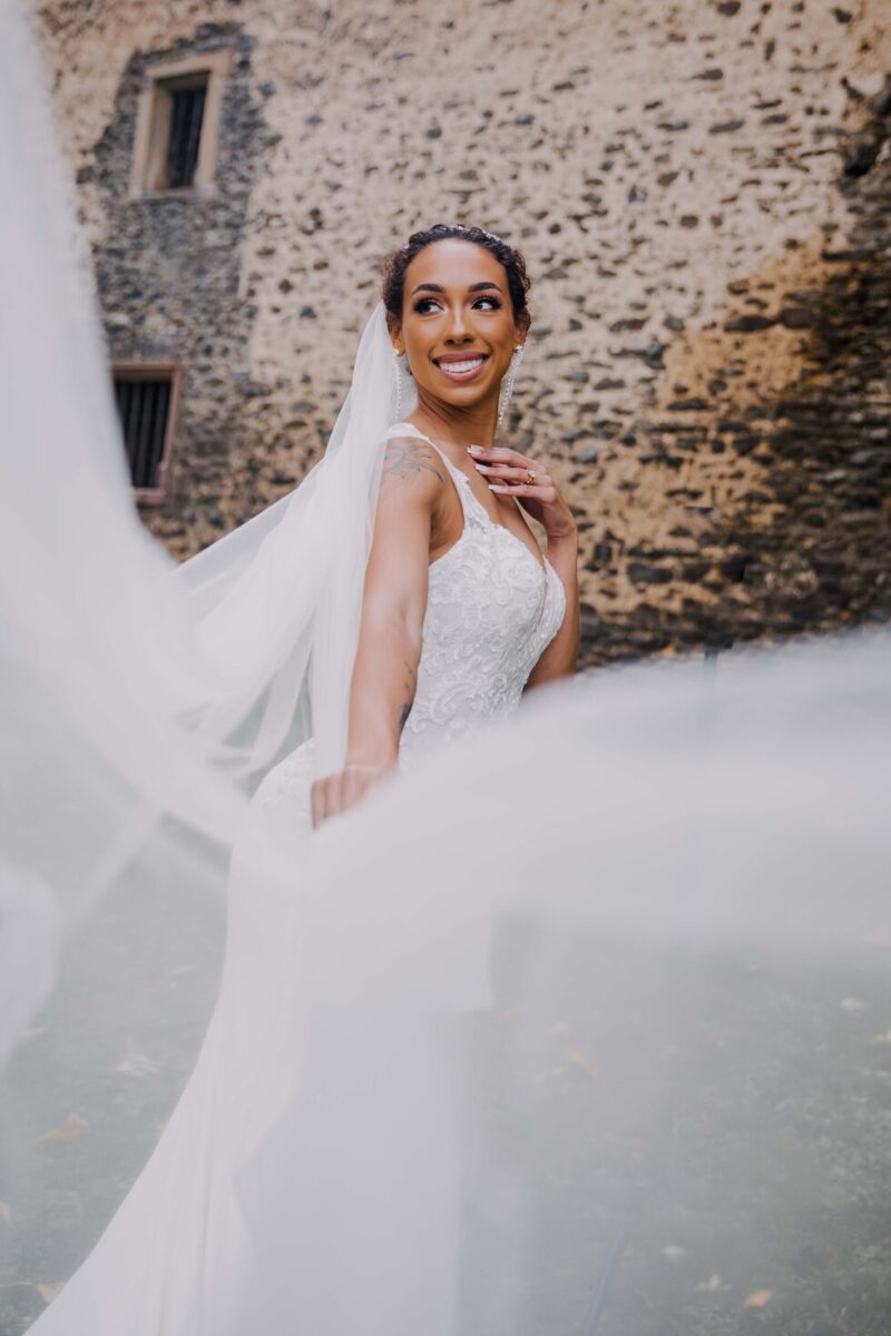 Eine fröhliche Braut steht im Freien an einer Steinmauer, strahlt und blickt zur Seite. Sie trägt ein weißes Brautkleid mit besticktem Mieder und einen langen Schleier, den sie zur Seite hält. Ihr Haar ist mit einem weißen Blumen-Accessoire hochgesteckt und fängt den Moment während des Fotoshootings perfekt ein. - Elis Wedding - Hochzeitsfotograf Frankfurt