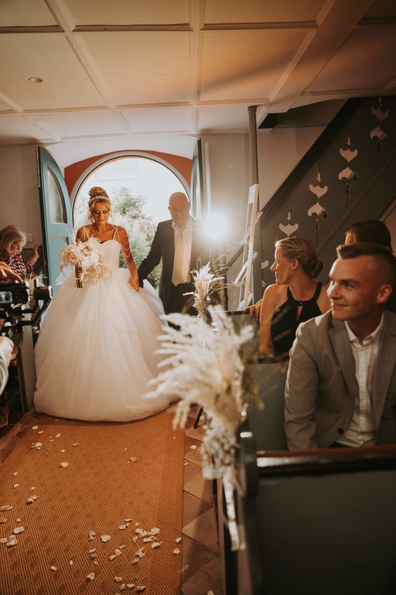 Eine Braut in einem voluminösen weißen Kleid und Schleier betritt einen sonnendurchfluteten Raum voller sitzender Gäste. Sie hält einen Blumenstrauß in der Hand und wird von einem Mann im schwarzen Anzug begleitet. Während Blütenblätter den Gang schmücken, fängt der Hochzeitsfotograf links jeden Moment ein. Die Gäste beobachten ihren Eintritt, einige lächeln und andere drehen sich auf ihren Sitzen um. - Elis Wedding - Hochzeitsfotograf Frankfurt