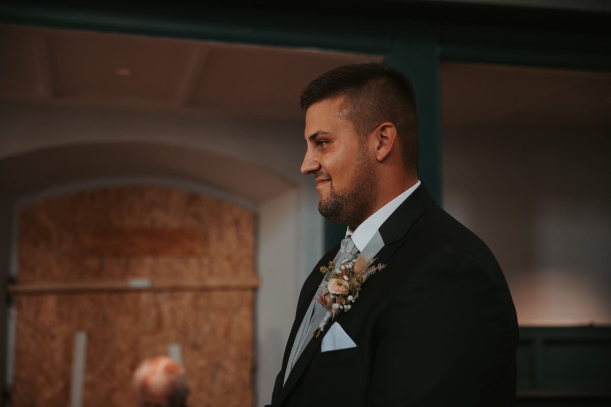 Ein Mann in dunklem Anzug, hellgrauer Krawatte und einem Blumenboutonniere steht in seiner Standesamtlichen Trauung in Frankfurt, lächelt und blickt nach rechts. Im Hintergrund sind ein grüner Torbogen und dahinter eine Holztafel mit sanfter, stimmungsvoller Beleuchtung zu sehen. - Elis Wedding - Hochzeitsfotograf Frankfurt